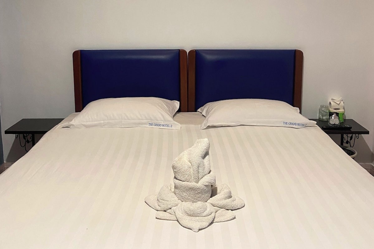 Una cama doble con toallas dispuestas, dos mesitas de noche, una lámpara de pared y dos cuadros enmarcados.