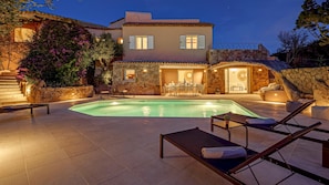 A poolside area with lounge chairs, a stone wall, and a building with a tiled roof.