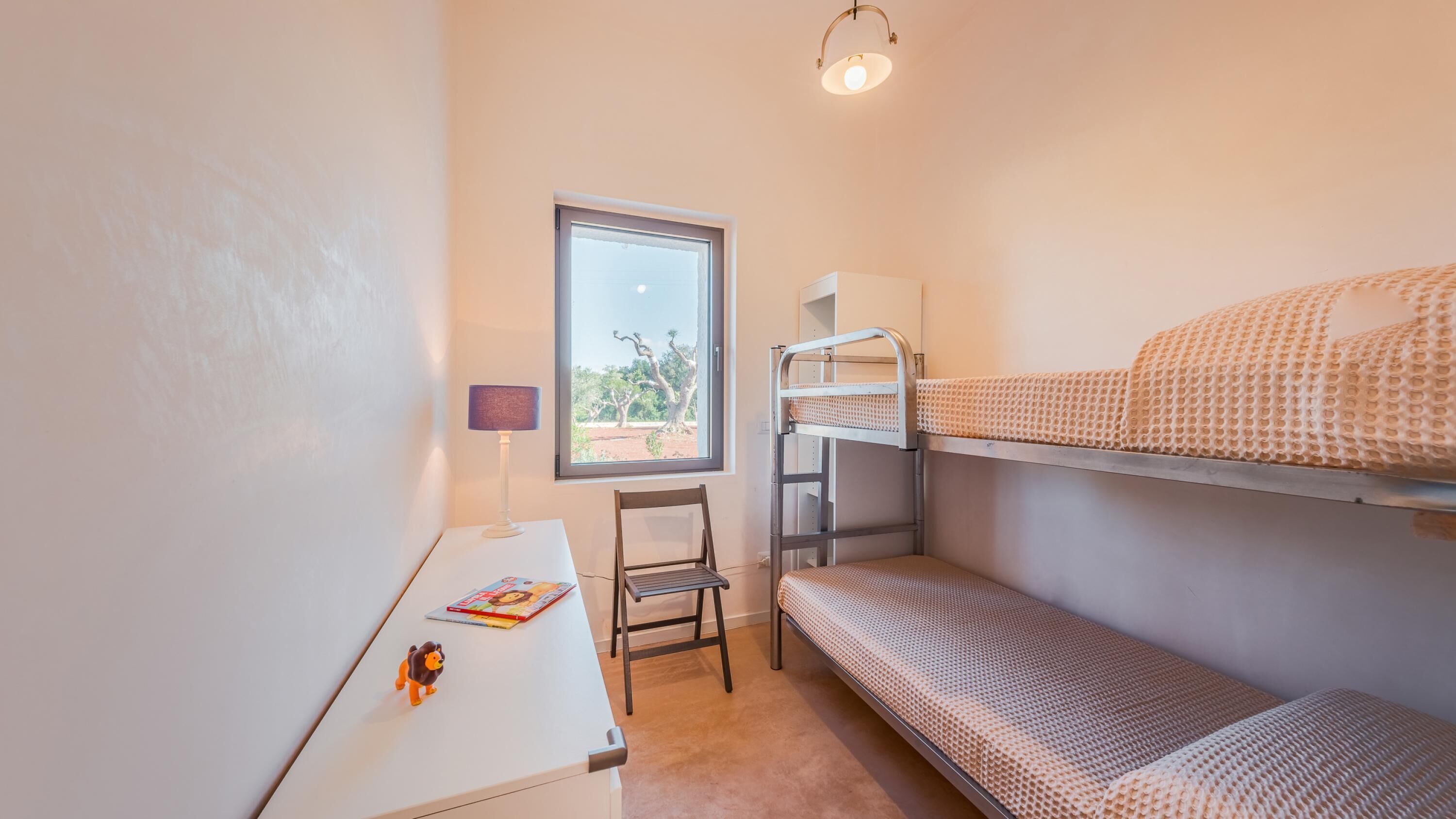 Ein kleines, kompaktes Zimmer mit Etagenbetten, einem Schreibtisch, einem Stuhl und einem Fenster mit Blick auf Bäume.