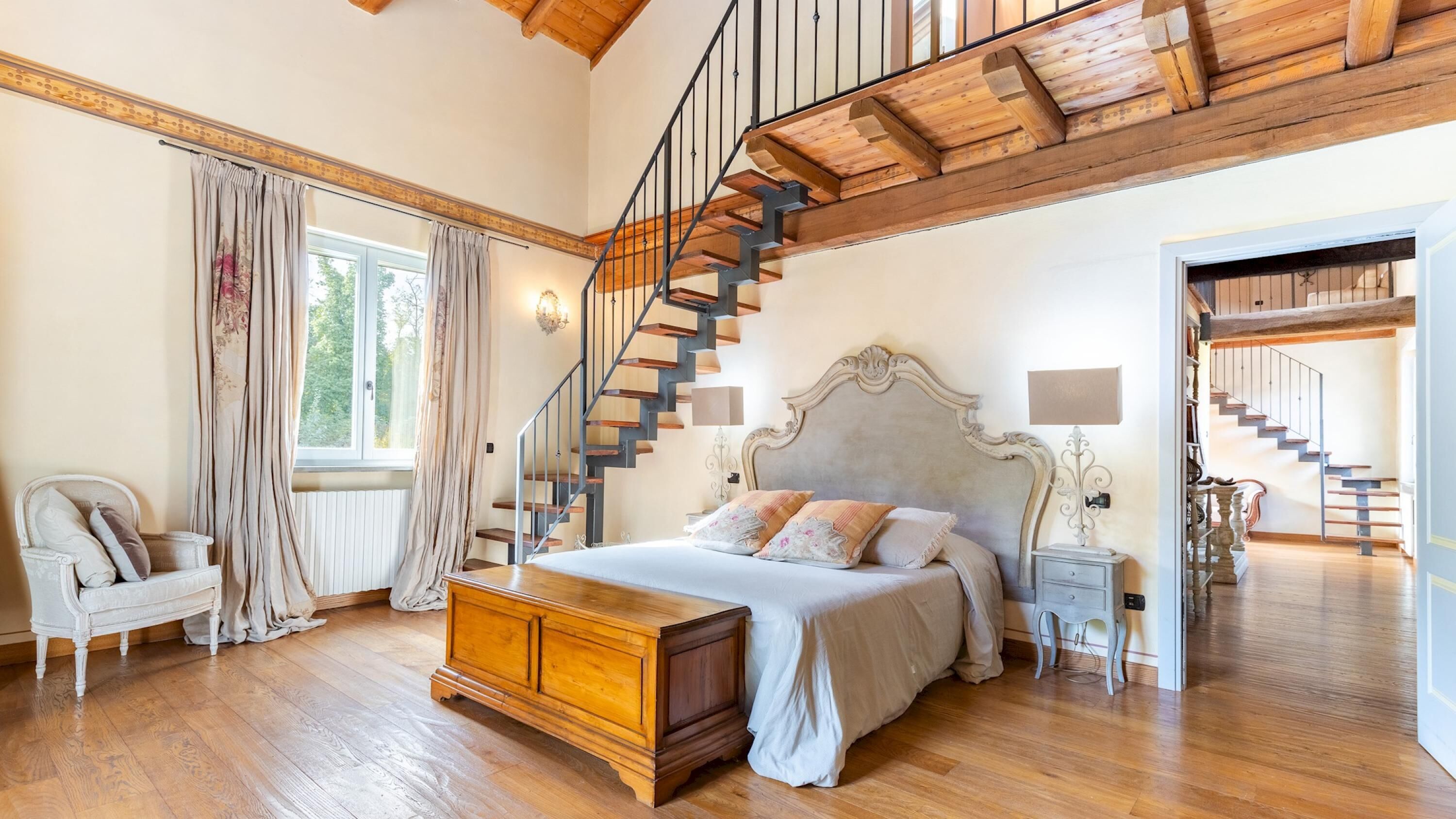 A spacious bedroom with a large bed, a wooden staircase, and a view of the outdoors.
