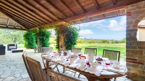 An outdoor dining area with a stone table set for a meal, surrounded by wooden chairs and benches, under a wooden pergola with greenery. - Cascina Moglia 8 1 Emma Villas (Carpeneto)