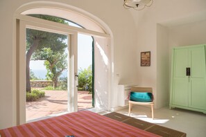 Une chambre à coucher comprenant un lit, une chaise, une armoire verte et une porte-fenêtre coulissante donnant sur un espace extérieur avec vue sur des arbres et un mur en pierre.