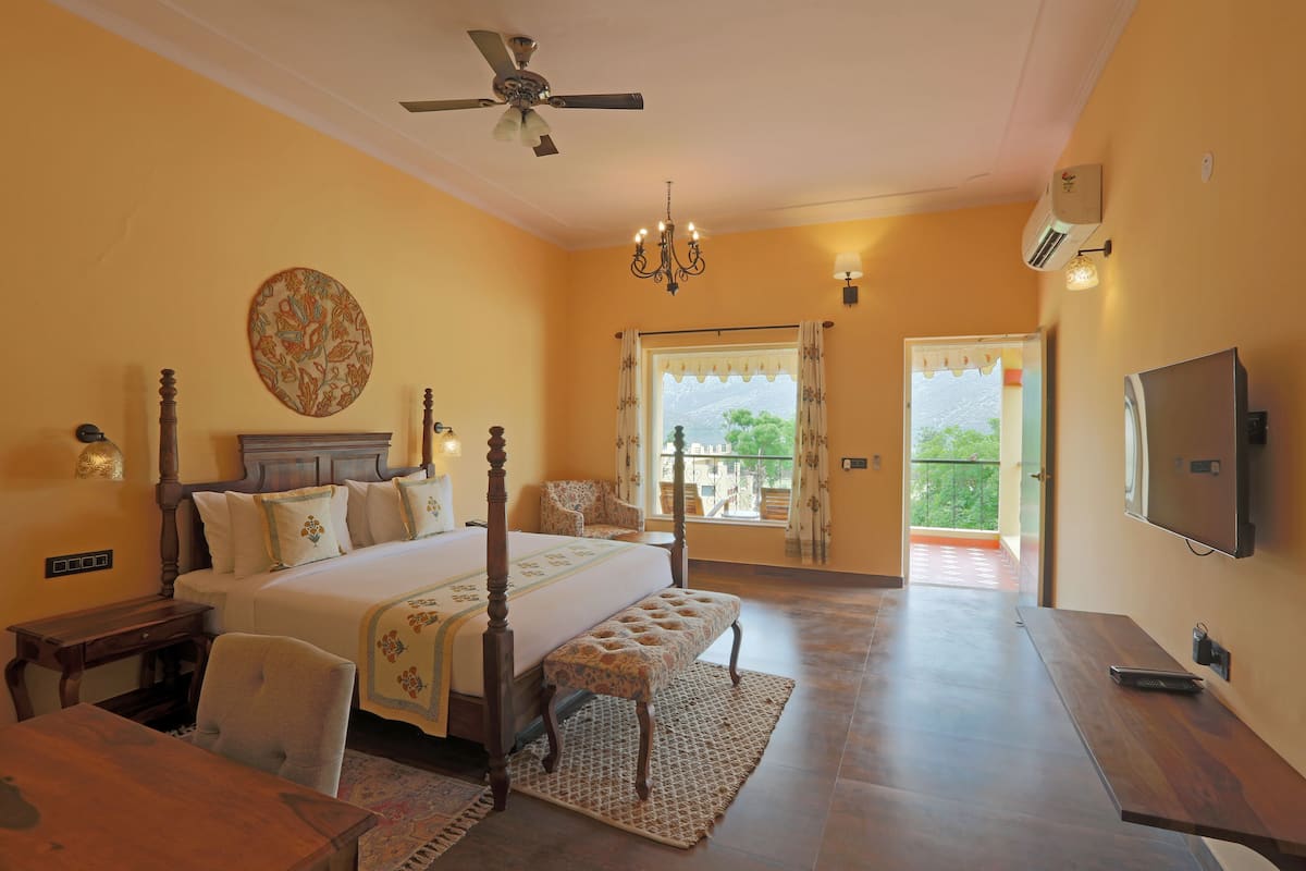 A bedroom with a four-poster bed, a desk, a chair, a bench, a chandelier, a ceiling fan, a TV, and a window with curtains.