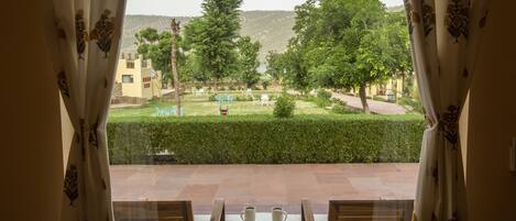 A view from a room with a table and chairs, overlooking a landscaped garden with a pool, trees, and a building in the background.