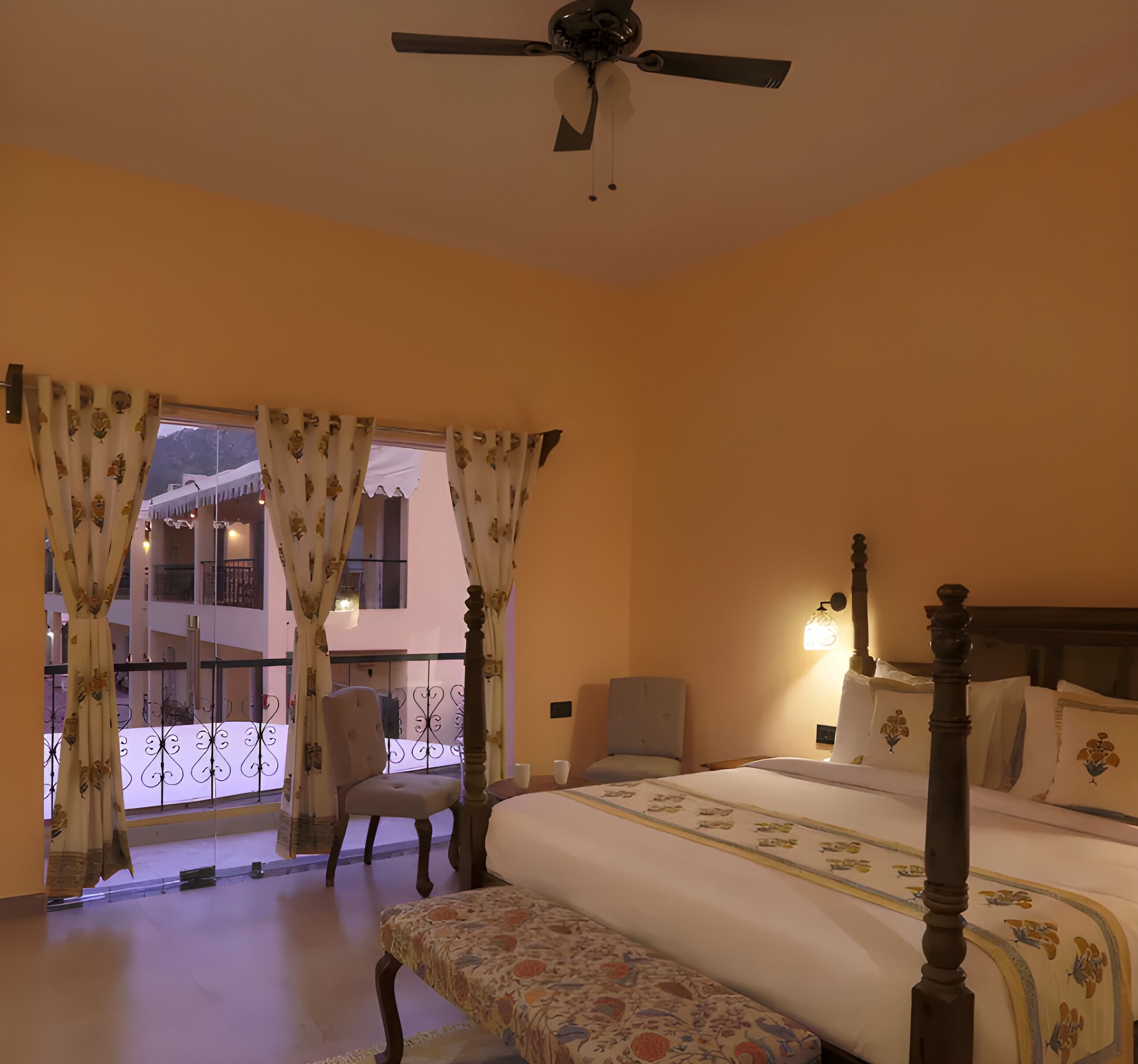 Quarto com cama, cadeira, mesa de cabeceira, ventoinha no teto e janela com cortinas.