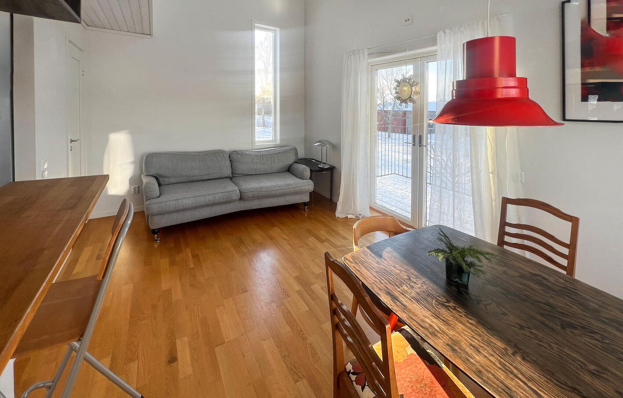 Una habitación con una mesa de comedor de madera, un sofá gris, una lámpara colgante roja y un ventanal con vistas a un paisaje nevado.