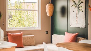 A cozy room with a wooden table, white chairs, a cushioned bench with pink pillows, a woven pendant light, and a window with a view of greenery. - luxury chalet on a cosy campsite near the sea (Westerland)