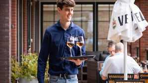 A person holding a tray with two glasses of wine, standing in front of a building with a brick facade and large windows. - luxury chalet on a cosy campsite near the sea (Westerland)