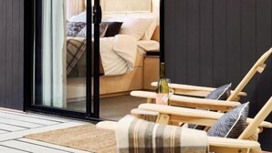A modern room with a sliding glass door leading to a bedroom, a wooden deck with lounge chairs, and a wall-mounted light fixture. - Southcamp (Dunsborough)