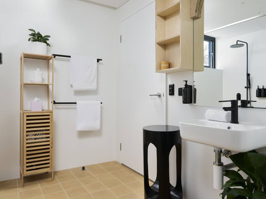 Un baño moderno con lavamanos blanco, pedestal negro, estante de madera y espejo sobre el lavamanos.