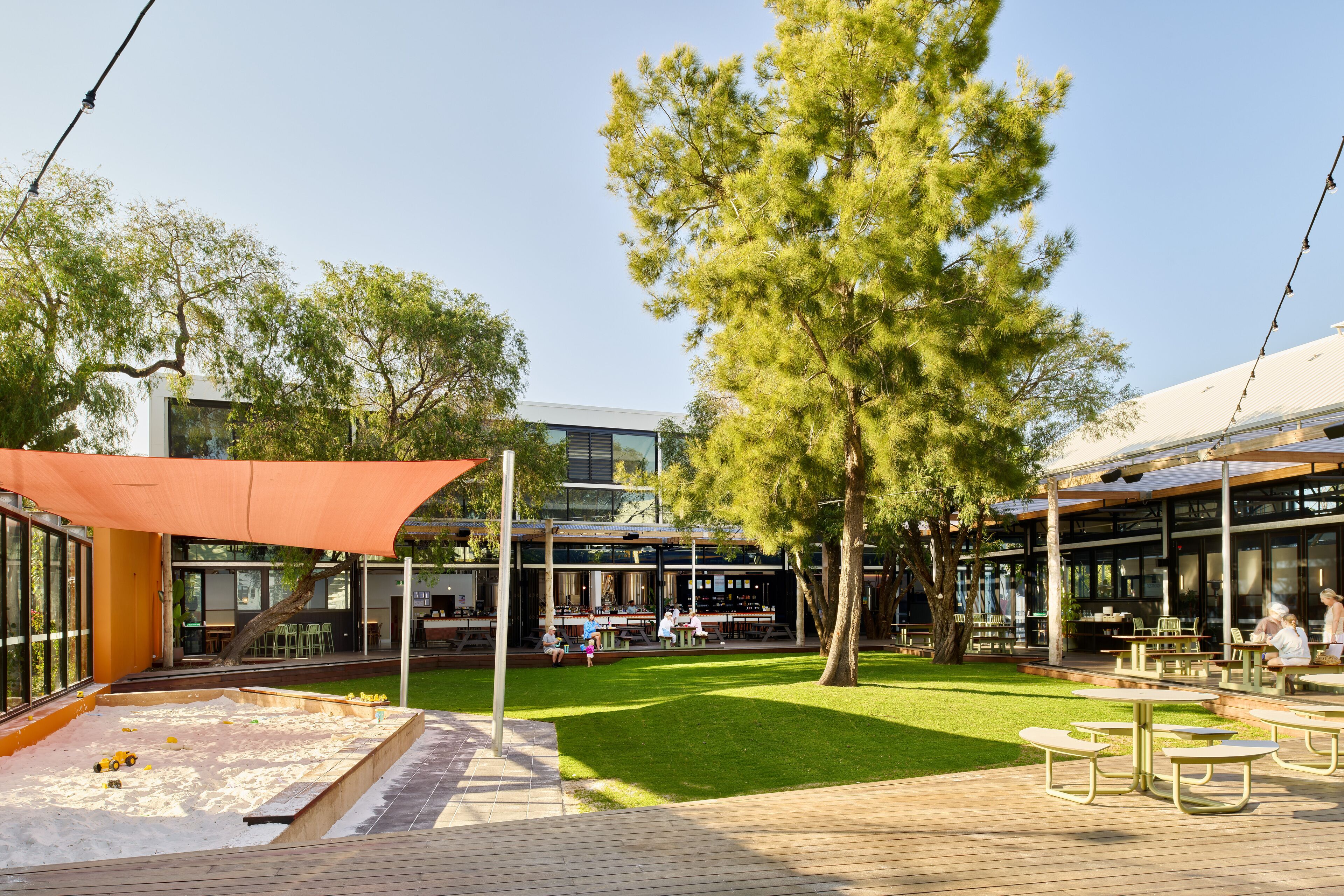 An outdoor patio area with tables and chairs, a sandpit, and a building with large windows and a covered seating area.