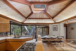 A modern kitchen with a central island, large windows, and a dining area with a long table. - Sea of Tranquility: Iconic Glass House in Forest Glade 3BR2BA 30Night min (Carmel)