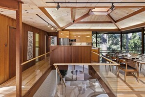 A modern kitchen with wooden cabinets, a stainless steel refrigerator, and a dining area with a glass table and chairs. - Sea of Tranquility: Iconic Glass House in Forest Glade 3BR2BA 30Night min (Carmel)