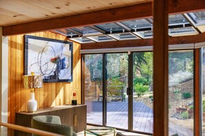 A modern interior with wooden paneling, a glass table, a framed abstract painting, and a view of an outdoor deck. - Sea of Tranquility: Iconic Glass House in Forest Glade 3BR2BA 30Night min (Carmel)