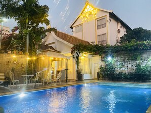 A swimming pool with a covered seating area, a building with a sign, and lush greenery. - Ananphada Boutique (Siem Reap)
