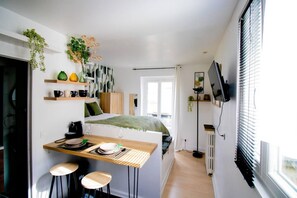 Un espace de vie compact comprenant un coin cuisine, un espace repas avec des tabourets, un lit recouvert d’une couverture verte et un téléviseur fixé au mur.
