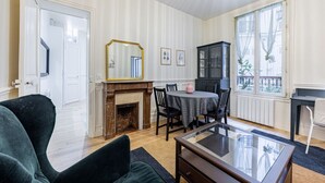 A room with a fireplace, a dining table with chairs, a glass coffee table, and a large window with a view of the outside. - Au cœur des Batignolles (Paris)