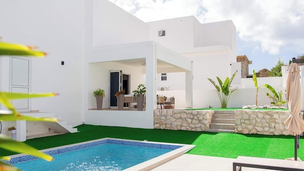 Una casa moderna con piscina, un patio con mesa y sillas, y un muro de piedra.