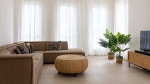 Una sala de estar moderna con un sofá modular, un taburete redondo tejido, una planta grande en maceta y un televisor sobre un mueble de madera.