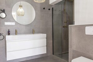 Un baño moderno con tocador blanco, cabina de ducha de vidrio y espejo redondo con lámpara colgante.