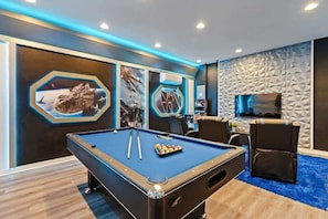 Una mesa de billar con superficie azul de fieltro, rodeada por una mesa de billar de color oscuro y una zona de estar con alfombra azul.