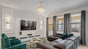 A modern living room with a sectional sofa, a green armchair, a flat-screen TV, a chandelier, and large windows with curtains. - 6BD Solterra Resort Near Disney (ST 5113) (Davenport)