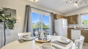 A dining area with a table set for four, a refrigerator, wooden cabinets, and a view of greenery through the window. - Mid-Good Day Venture (Colorado Springs)