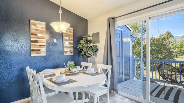 A dining area with a round table and white chairs, a hanging light fixture, and a view of the outdoors through a sliding glass door. - Mid-Good Day Venture (Colorado Springs)