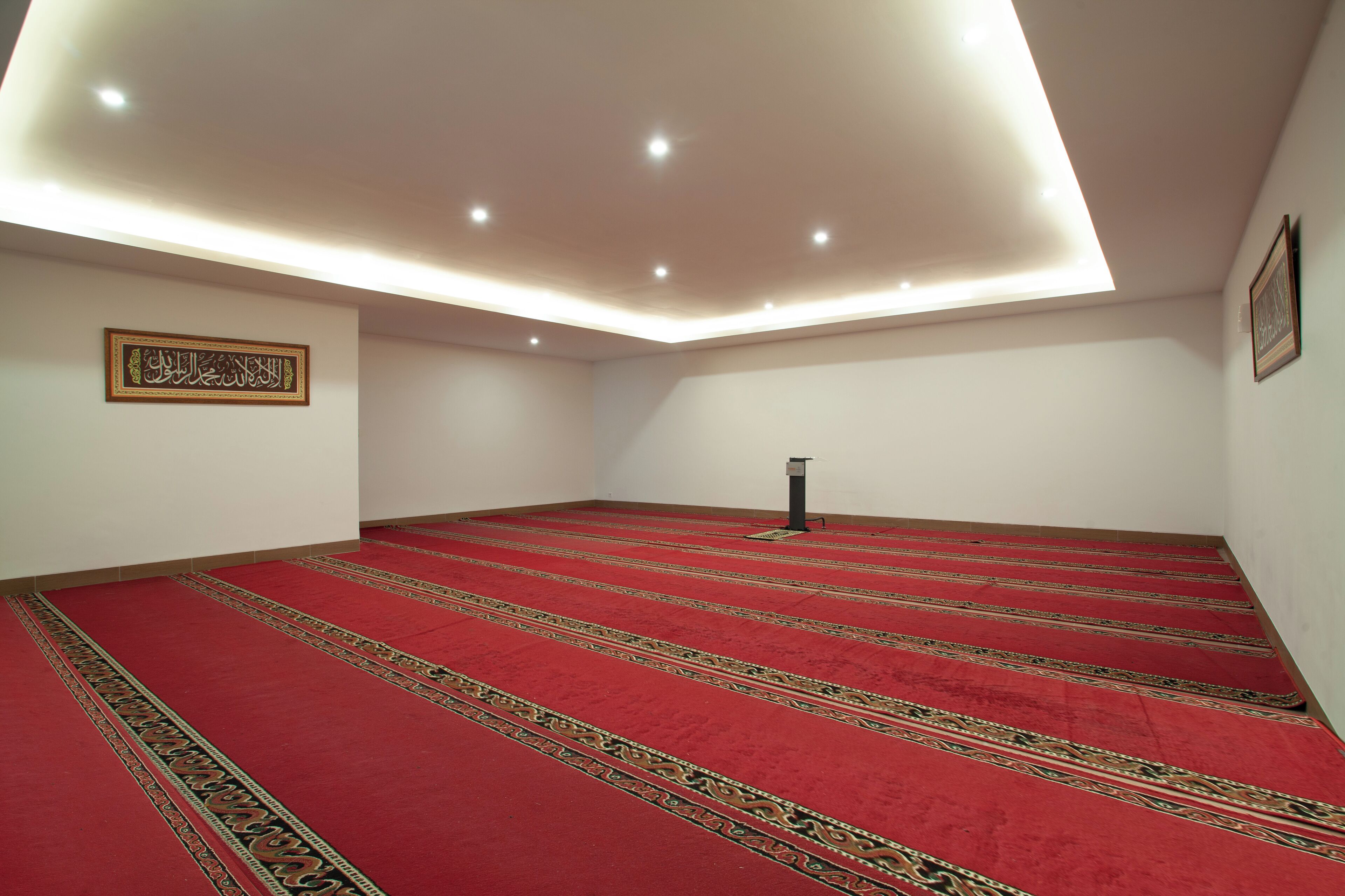 A room with a red carpet and a prayer mat, a ceiling light fixture, and a framed Arabic calligraphy on the wall.
