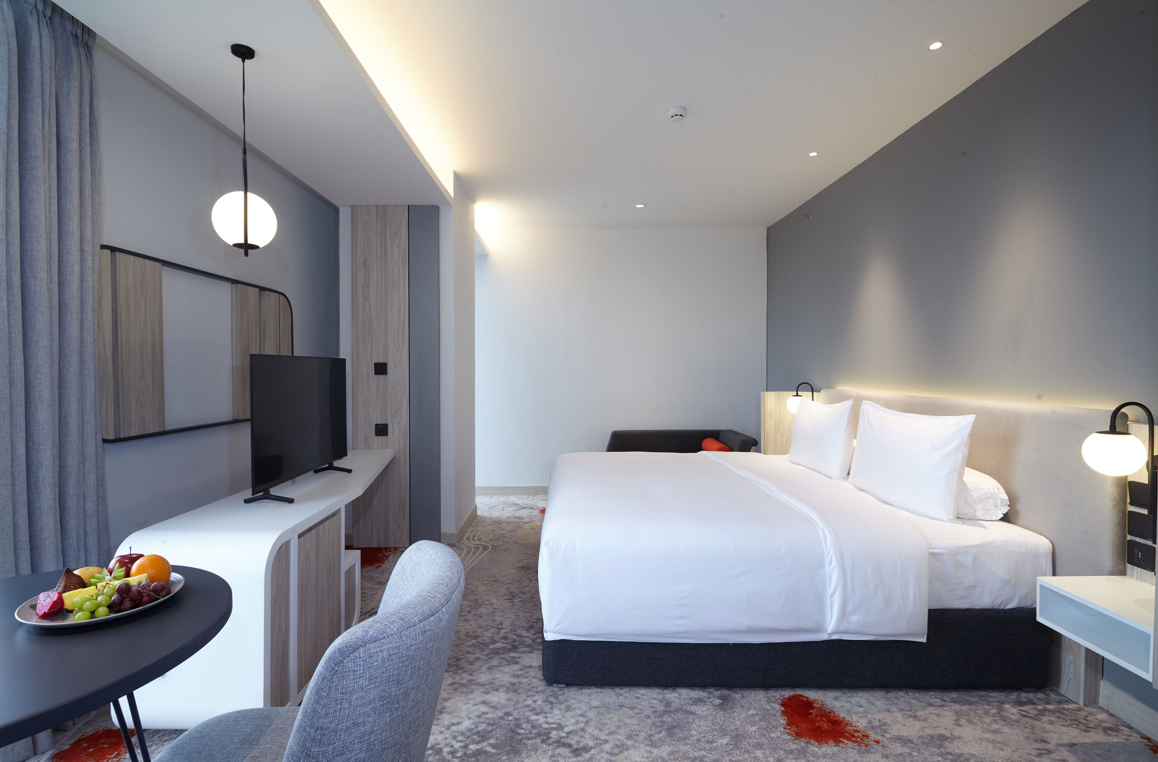 Una habitación de hotel moderna con cama, mesita de noche, televisión y un pequeño escritorio con una bandeja de frutas.