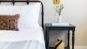 A neatly made bed with a patterned pillow, a bedside table with a book and a vase of flowers, and a woven basket on the floor.