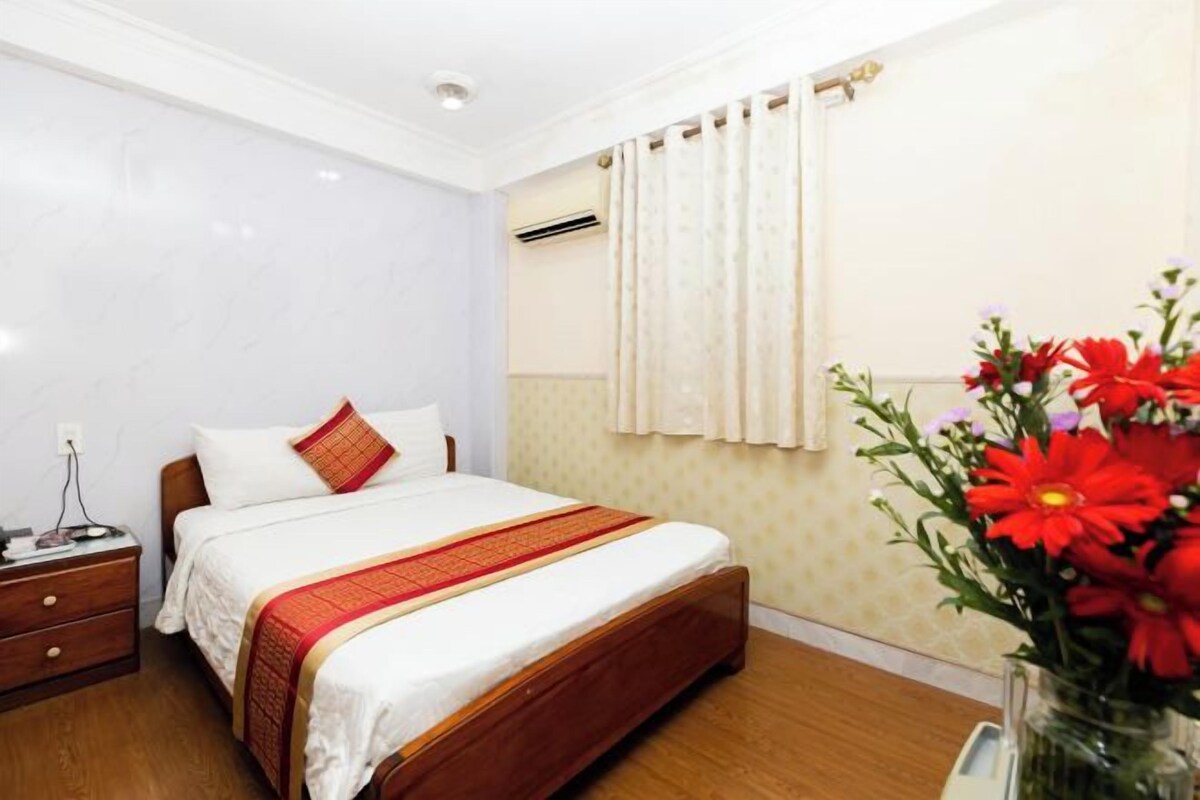 A hotel room with a bed, a nightstand, a vase of flowers, and a window with curtains.