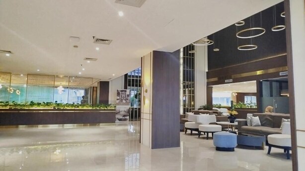 Un vestíbulo amplio con diseño moderno, que incluye una zona de estar con sofás y una mesa de centro, un mostrador de recepción y un gran ventanal con vistas al exterior.