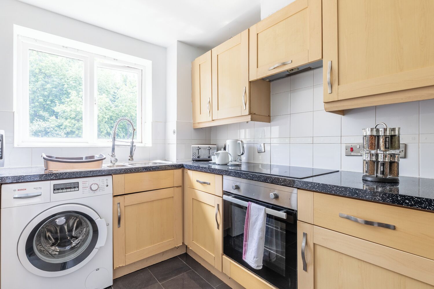 A modern kitchen with wooden cabinets, a Bosch washing machine, a built-in oven, and a window with a view of greenery.