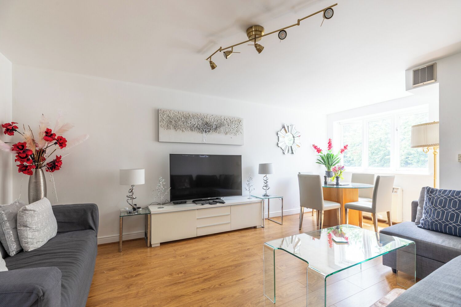 A modern living room with a flat-screen TV, grey sofas, a glass coffee table, and a dining area with a glass table and chairs.