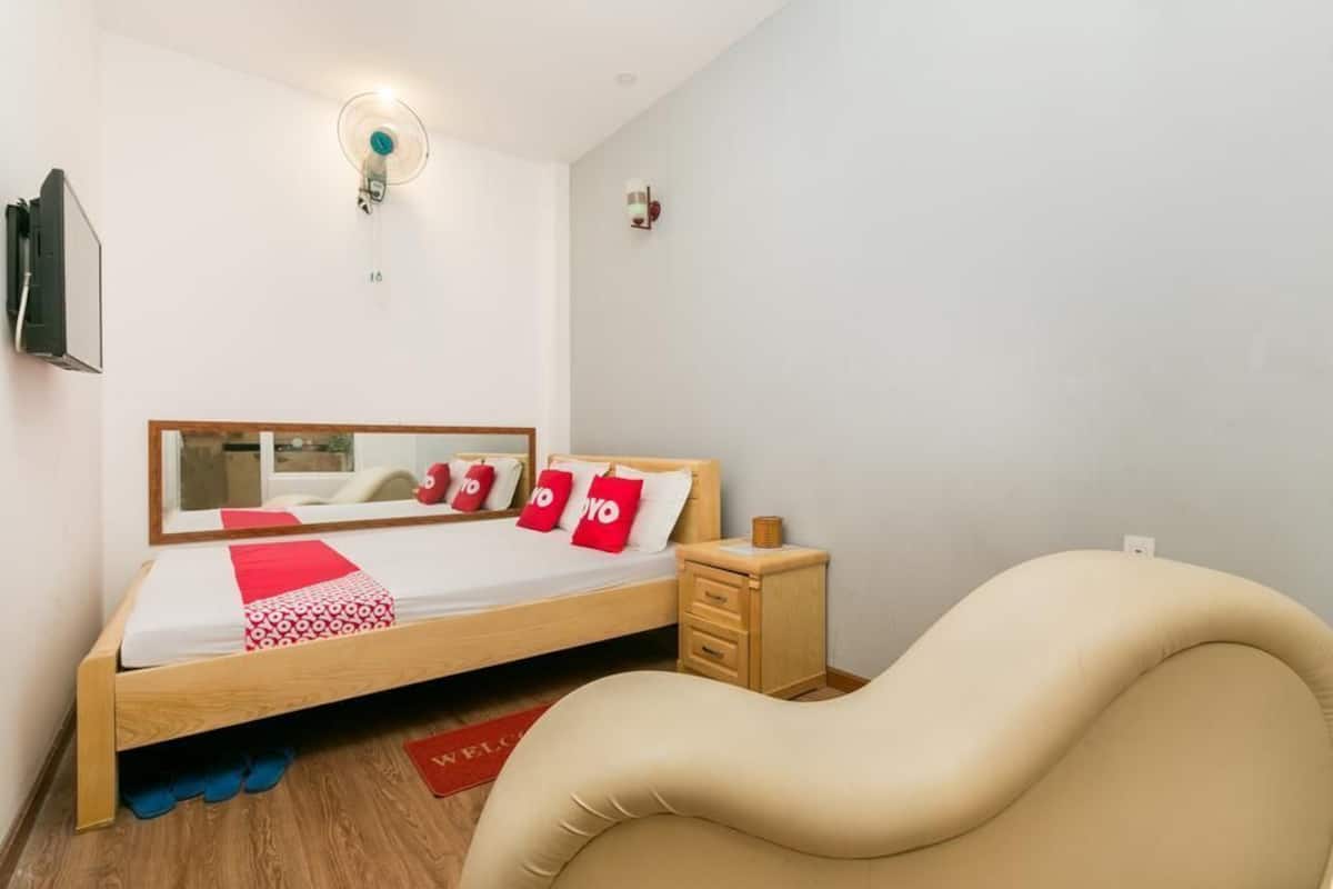 Une chambre à coucher avec un lit, une table de chevet, un miroir, un ventilateur fixé au mur et un paillasson rouge.