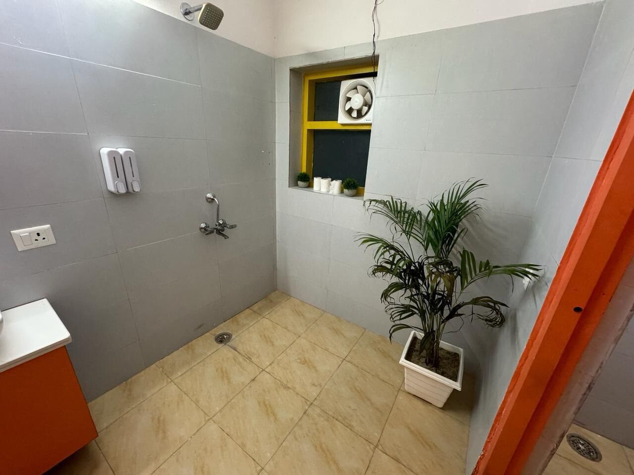 Un baño pequeño con baldosas grises, ducha, ventana con marco amarillo y una planta en maceta.