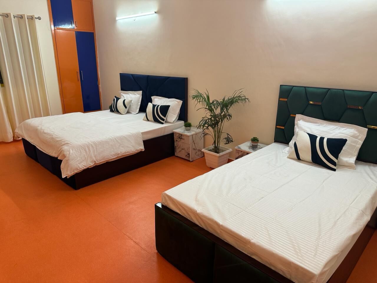 A hotel room with two beds, each with a headboard, white bedding, and decorative pillows. There is a small table with a plant and a nightstand with a lamp.