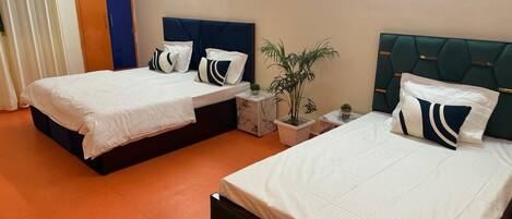A hotel room with two beds, each with a headboard, white bedding, and decorative pillows. There is a small table with a plant and a nightstand with a lamp.