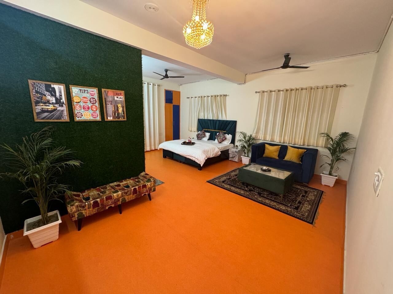 A room with a bed, a sofa, a coffee table, and a small bench. There are potted plants and a wall with framed pictures.