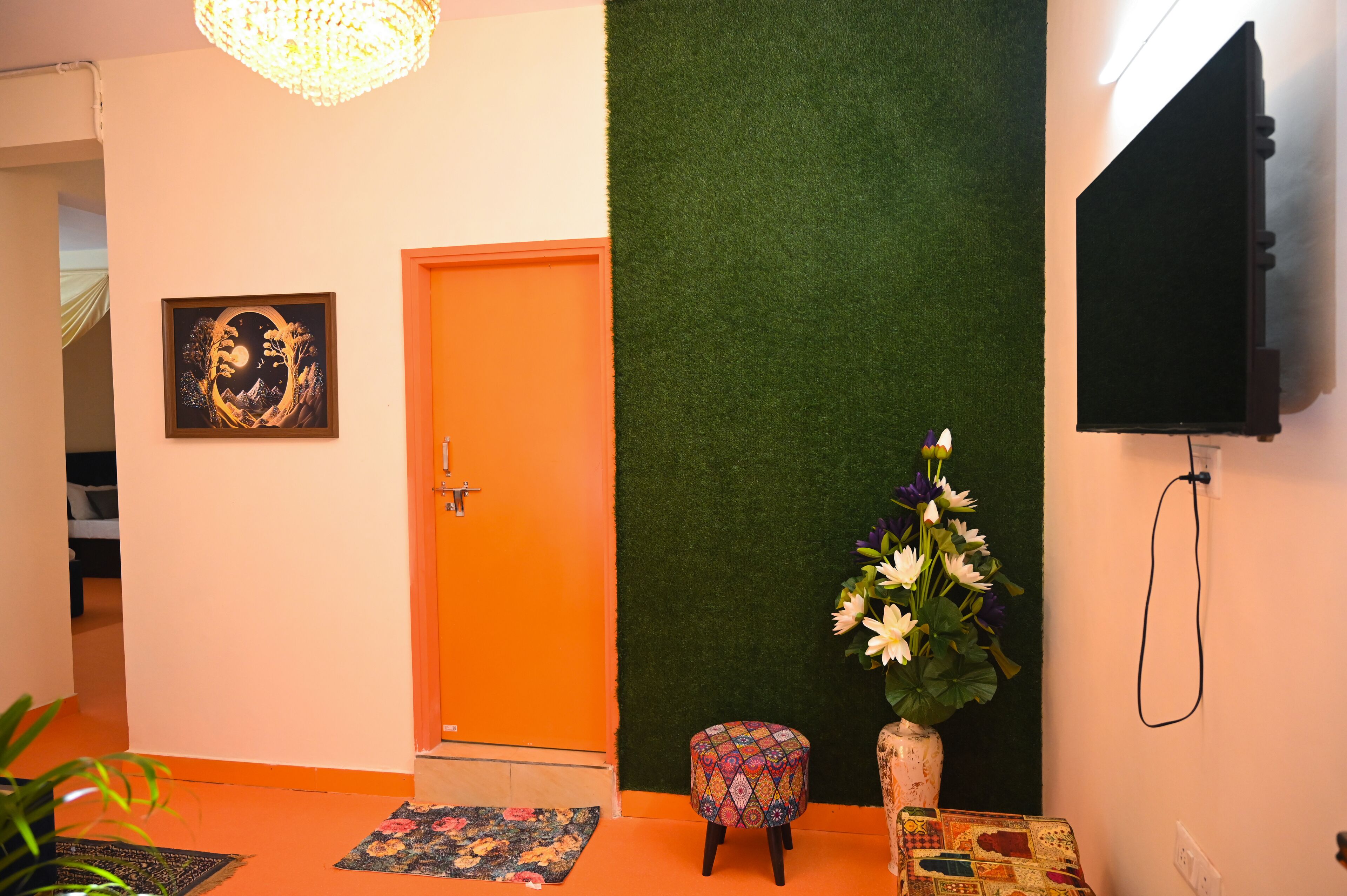 A hallway with an orange door, a framed artwork, a small stool, a vase with flowers, and a television mounted on the wall.