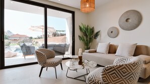 A modern living room with a sofa, patterned chairs, a coffee table, and a large window with a view of a pool and buildings. - Melior Luxury Villas Preveza (Preveza)