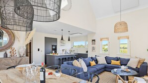 A modern living room with a large sectional sofa, a dining table with wine glasses and a bottle, and a kitchen area with a bar. - 3 The Vista, Willingcott Nr Woolacombe - Hot tub/ Pet friendly/ On-site pool/ EV Charger/ Ecohome (Woolacombe)