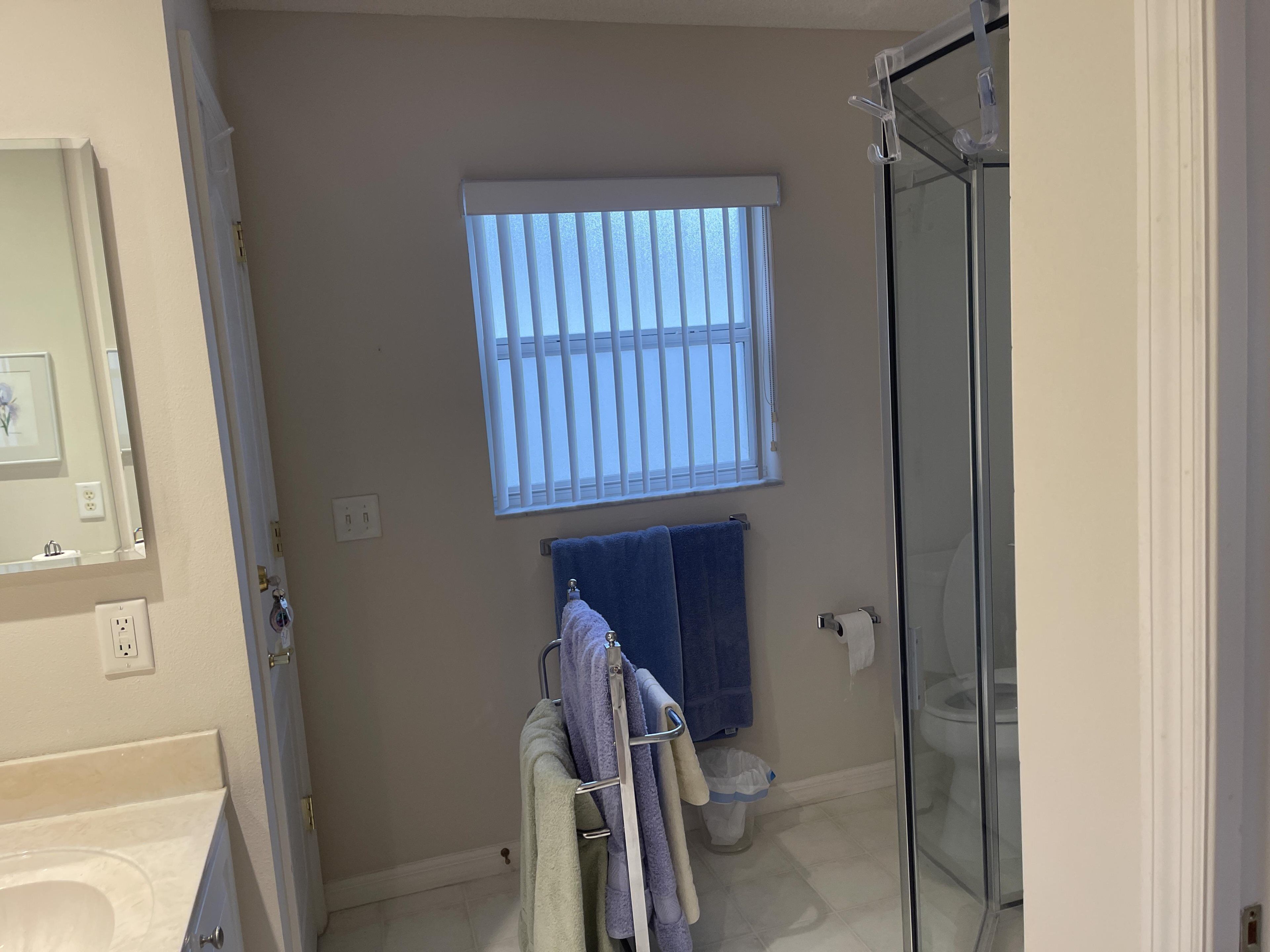 Une salle de bain comprenant des toilettes, un lavabo, un miroir, une fenêtre avec des stores et un espace douche.