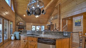 A wooden cabin interior with a kitchen area featuring granite countertops, a dishwasher, and a stone fireplace. - Stokes Haven - Fireplace & Hot Tub - Pet Friendly (McCaysville)