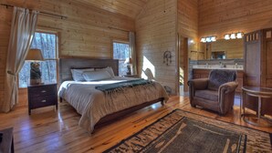 A cozy log cabin bedroom with a bed, bedside table, lamp, chair, and a view of the kitchen. - Stokes Haven - Fireplace & Hot Tub - Pet Friendly (McCaysville)