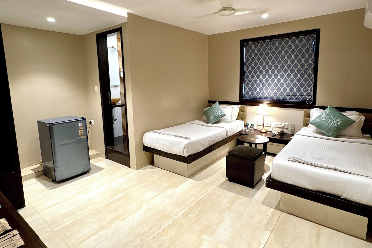 A hotel room with two beds, a small table, a refrigerator, and a window with a patterned blind.