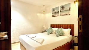 Una cama bien hecha con almohadas, una lámpara de noche y un cuadro de palmeras en la pared.