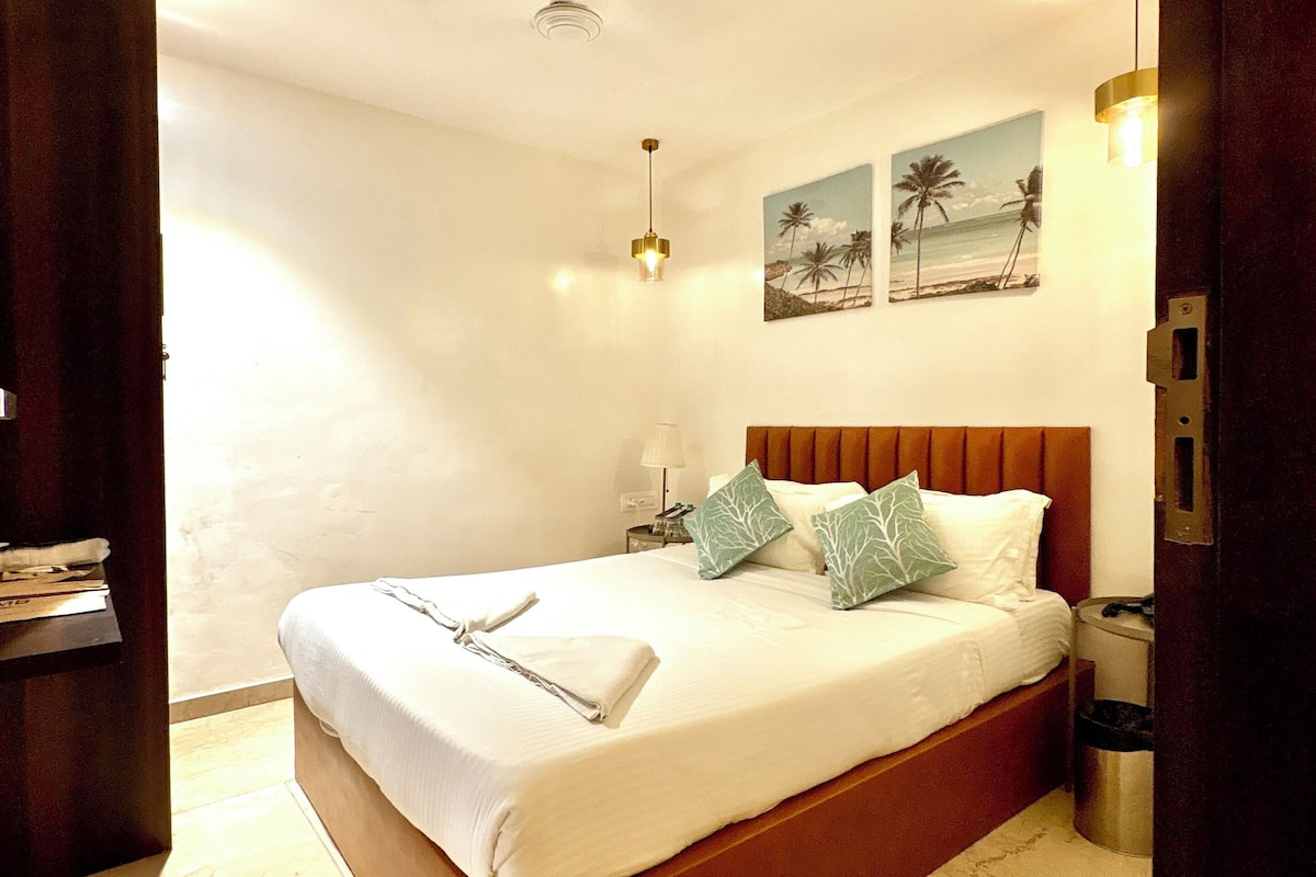 A neatly made bed with pillows, a bedside lamp, and a painting of palm trees on the wall.