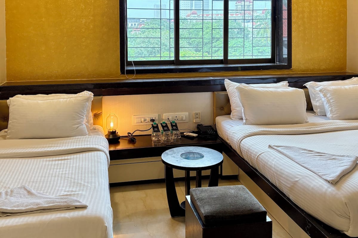 A hotel room with two beds, a small table, a window with a view of a building and greenery, and a ceiling fan.
